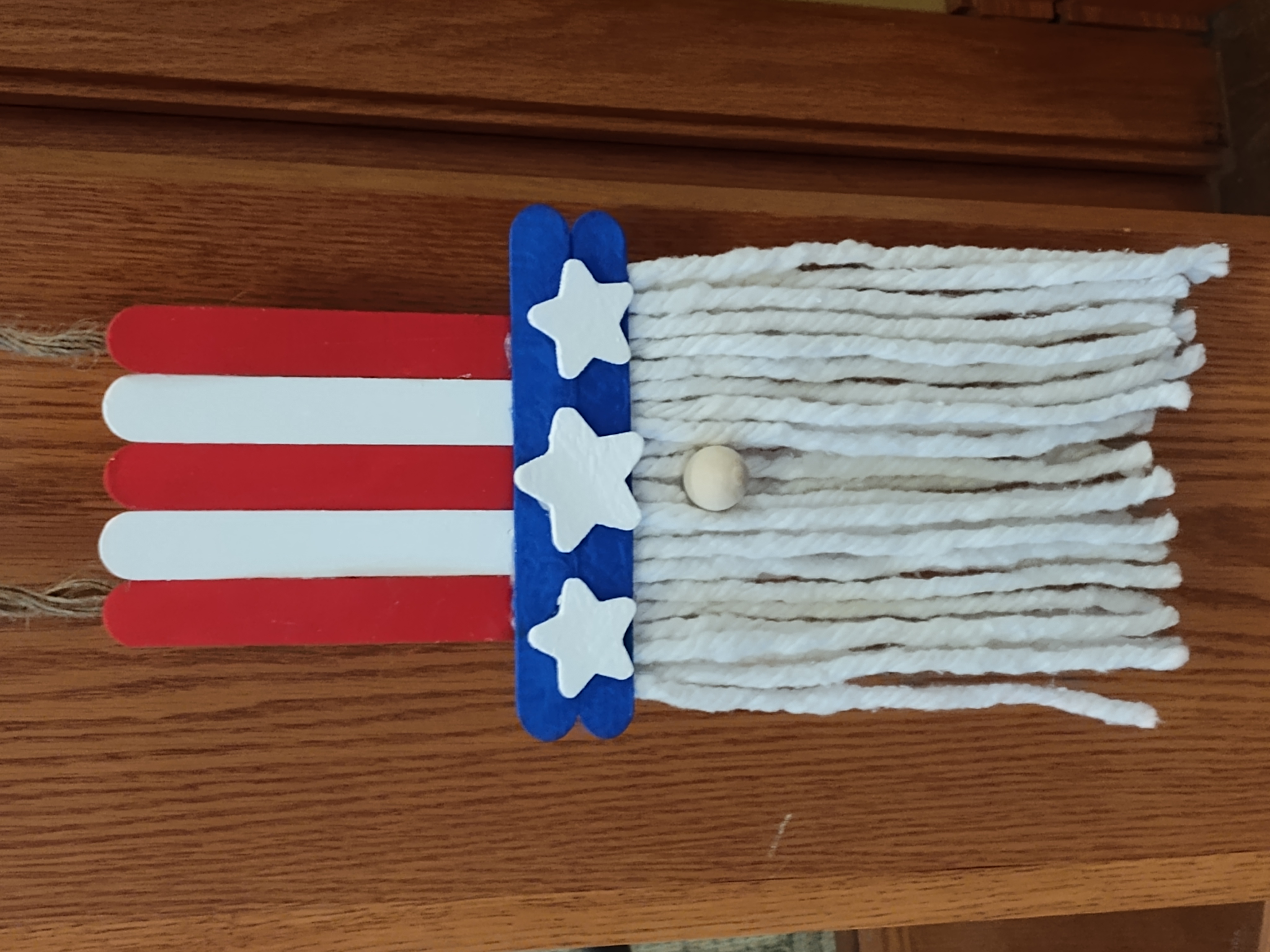 craft stick red, white, and blue top hat with white stars on the brim a white beard and a nose making an Uncle Sam gnome with a jute hanger.