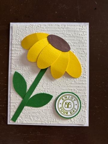a card with a white background on which a brown eyed daisy made of paper sticks out. The petals are yellow, center is brown, and leaf and stem are green. A small green stamp is on the side saying 'enjoy your day".